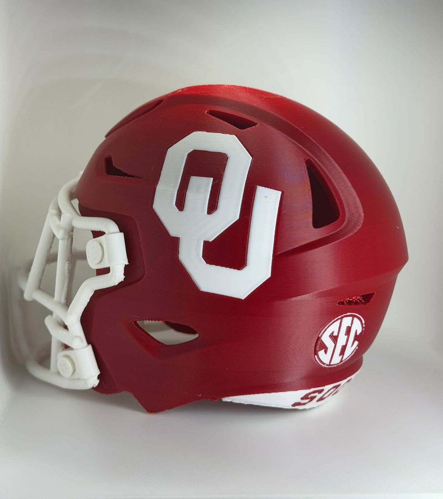 Side view of a miniature red football helmet with a white OU logo, a white face mask and an SEC decal